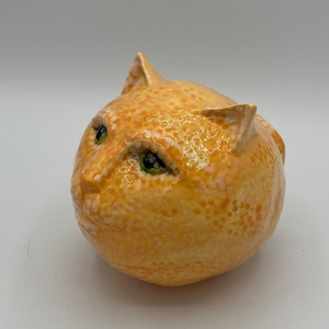 May include: A ceramic sculpture of a cat with a yellow and orange glaze. The cat has green eyes and a round, plump body.