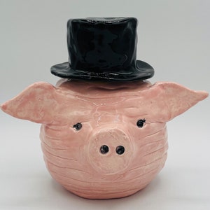 May include: A pink ceramic pig figurine wearing a black top hat. The pig has a detailed snout and ears.