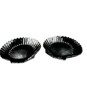 Puede incluir: Dos platos negros en forma de concha con un acabado brillante. Cada plato tiene un borde festoneado y un interior texturizado y acanalado. Los platos se muestran sobre un fondo blanco, resaltando su color oscuro y su forma única.