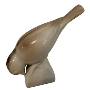 Puede incluir: Figura de pájaro de cerámica en tonos marrón y crema. El pájaro está representado en una pose elegante, con la cabeza inclinada y el pico apuntando hacia abajo. La figura tiene un acabado brillante y está posada sobre una pequeña base.