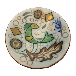 May include: A small, round ceramic plate with a hand-painted design. The plate features a green bird with a yellow beak and a brown leaf. Gold and brown accents and geometric shapes decorate the white background. The rim is brown.