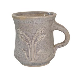 May include: A ceramic mug with a speckled, muted grey and brown glaze. The exterior features an embossed design of three stalks of wheat centered on the front. The mug has a wide, flared rim and a sturdy, curved handle on the right side.