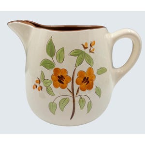 Puede incluir: Jarra de cerámica color crema con borde y asa marrones. La jarra está decorada con flores naranjas y marrones, hojas verdes y pequeñas bayas naranjas. Tiene un pico vertedor.