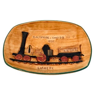 May include: A tan ceramic dish with a green rim featuring a raised relief of a black and gold train with red wheels. The text "Baltimore & Ohio R.R. 1837" and "Lafayette" are also on the dish.