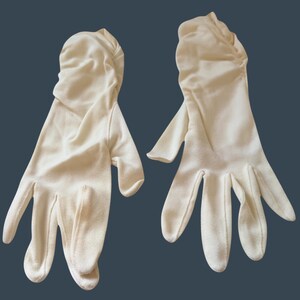 Peut inclure: Paire de gants blanc cassé, probablement pour une tenue de cérémonie, sur fond bleu foncé. Les gants sont en tissu doux, avec un détail froncé au poignet. Les doigts sont bien définis.