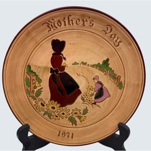 May include: A decorative plate with a Mother's Day theme, featuring a woman and child in vintage attire, surrounded by flowers and a fence. The plate is tan with a dark red rim and the year 1971.
