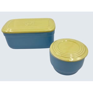 May include: Two-piece ceramic set in blue and yellow. The rectangular container has a yellow lid with a raised emblem. The round container has a yellow lid with concentric rings. Both pieces are on a white background.