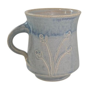 May include: A ceramic mug with a light blue and grey speckled glaze, featuring a flared rim and a curved handle. The exterior surface is decorated with a subtle, incised line drawing of three slender floral stems with small circular buds.