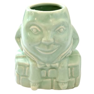 Puede incluir: Una maceta de cerámica verde claro con forma de cara sonriente. La maceta tiene una abertura redonda en la parte superior y está decorada con detalles como una pajarita y un patrón de ladrillos en la base.