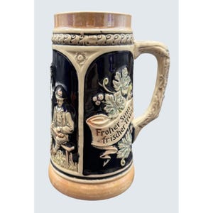 May include: A ceramic beer stein featuring a dark blue body with cream-colored relief accents. The design includes a seated figure, grape vine motifs, and a banner with the text Froher Sinn und frischer Trunk. It has a textured handle.