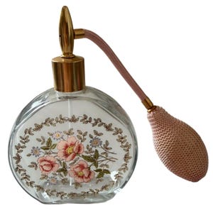 May include: A clear glass perfume bottle with a gold-tone metal atomizer pump and a pink fabric-covered bulb. The front of the bottle features a decorative oval border of green leaves surrounding a central cluster of pink and blue floral illustrations.