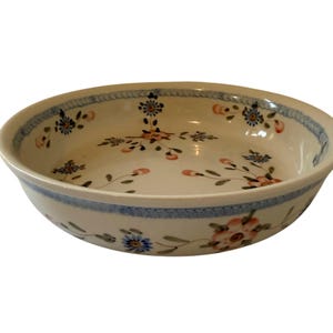 May include: A round ceramic bowl with a cream-colored base featuring hand-painted floral motifs in shades of blue, orange, and green. The rim is decorated with a textured blue band, and the interior displays delicate sprigs of flowers.