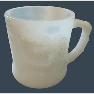 May include: A white milk glass mug featuring a translucent, milky finish and a textured, embossed design on the exterior surface. The mug has a rounded body, a wide circular rim, and a curved handle extending from the side of the vessel.