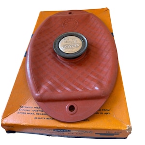 May include: A vintage red rubber ice cap with a screw-on metal cap, resting on its original orange cardboard box. The cap is marked "ONE TURN SEALS IT" and "DAVOL". The box has text including "STANDARD ICE CAP".