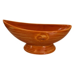 May include: An orange ceramic boat-shaped bowl with a pedestal base. The bowl has a textured, wood-grain appearance and a small, decorative floral element on the front. The bowl is a warm, earthy tone.