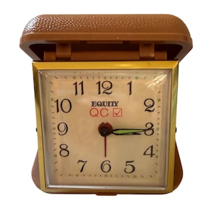 May include: A square travel alarm clock with a brown textured plastic case that opens on a hinge. The clock face is off-white with black numerals, a gold-tone metal frame, and the text Equity QC with a red checkmark symbol in the center.