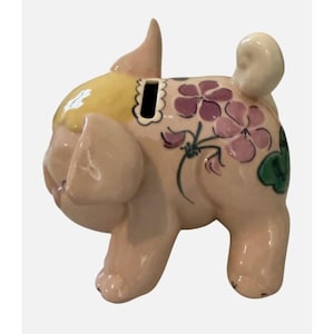 May include: A ceramic pug-shaped piggy bank in a light pink hue, adorned with purple floral designs and green leaves. The pug's head is yellow, and it has a slot for coins on its back. The bank has a glossy finish.