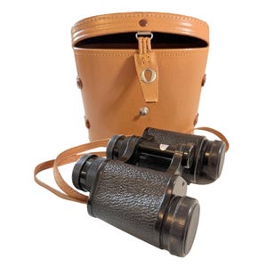 Op de afbeelding: Zwarte verrekijker met een getextureerde grip en een centraal scherpstelwiel, vergezeld van een beige lederen draagtas met een klepsluiting met metalen drukknoop en een bijpassende beige lederen nekriem voor draagbaarheid.