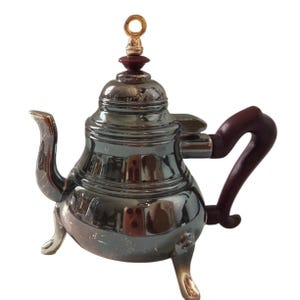 Puede incluir: Una tetera plateada brillante con un asa y detalles en marrón oscuro. La tetera tiene un diseño decorativo y se apoya en tres patas pequeñas. La tapa está rematada con un anillo dorado y un adorno marrón oscuro.