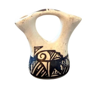 Puede incluir: Un jarrón de cerámica blanco con un asa grande y un diseño geométrico azul oscuro alrededor de la base. El jarrón tiene una forma única con una abertura ancha y un asa curva. El diseño incluye espirales, líneas y formas.