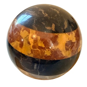 Puede incluir: Una esfera decorativa con superficie pulida. La esfera presenta bandas de colores negro, marrón y beige. Las bandas están dispuestas horizontalmente alrededor de la esfera. La esfera es un objeto decorativo.