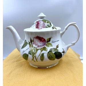 May include: White porcelain teapot with a lid, decorated with a rose design in pink and green. The teapot has a curved spout, a loop handle, and a gold rim. The words "Rose mossa" are printed on the teapot.