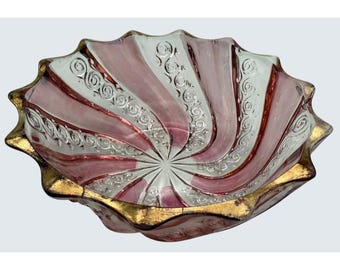 May include: A decorative glass bowl with a spiral design. The bowl has alternating pink and white stripes, with silver scrollwork. The rim is edged with gold, adding elegance to this unique piece. A stylish addition to any home.