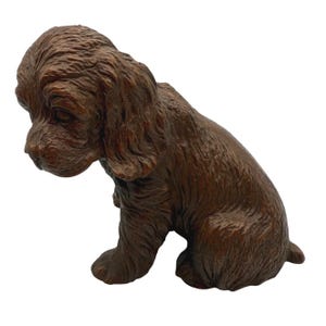 May include: A detailed, brown sculpture of a puppy in a seated position, with its head tilted downward. The puppy has long, floppy ears and a textured coat, giving it a realistic appearance. The sculpture is set against a plain white background.