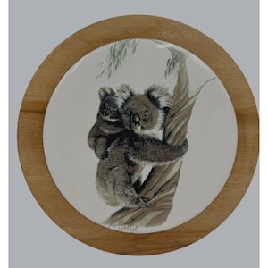 May include: Circular wooden tray with a white ceramic centre showing a detailed illustration of two koalas on a tree. The koalas are in grey and brown, with a light brown tree trunk. The tray has a wooden frame.