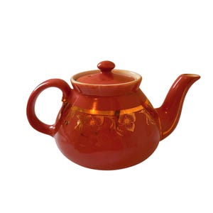 Puede incluir: Una tetera de cerámica redonda y brillante de color naranja quemado con una banda dorada en el centro. La tetera está decorada con un delicado patrón floral dorado repetitivo e incluye una tapa a juego con un pequeño asa redondeada.
