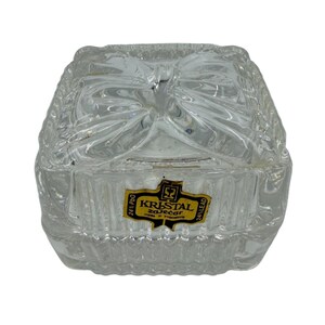 May include: Clear lead crystal trinket box with a bow-shaped design on the lid. The box has a square shape with textured sides and a label that reads "Kristal Zaječar".