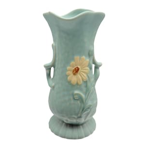 May include: A light blue ceramic vase with a textured surface, featuring two side handles and a raised, cream-colored daisy flower design on the front. The vase has a scalloped rim and a wide, ribbed base, showcasing a glossy finish throughout.