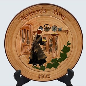 May include: A ceramic plate in shades of brown and tan, featuring a relief design of a kitchen scene with a woman, the words "Mother's Day", and the year "1975". The plate is displayed on a black stand. The design has a vintage feel.