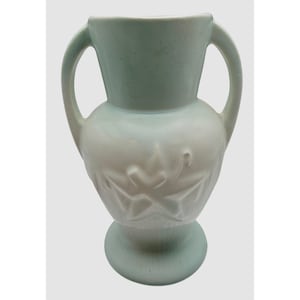 May include: A pale mint green ceramic vase featuring two symmetrical side handles, a wide flared rim, and a rounded body decorated with a raised, embossed star pattern. The base is circular and provides a stable foundation for the vessel.