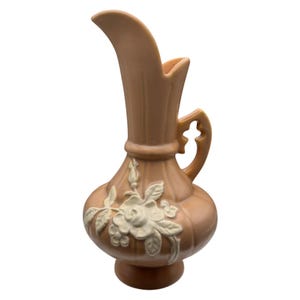 May include: A peach-coloured ceramic pitcher featuring a tall, fluted neck and a handle with a decorative cutout. The bulbous body is adorned with a raised, white floral relief pattern depicting roses and leaves, set against a smooth, glossy finish.