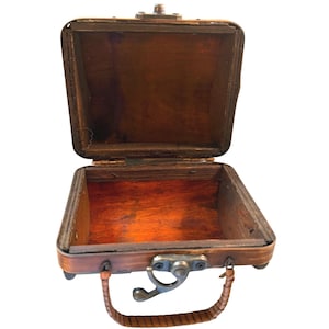 May include: A small, open wooden box with a hinged lid and a dark brown finish. The box features a metal latch on the front and a curved, textured handle attached to the base. The interior is hollow with visible wood grain and dark brown tones.