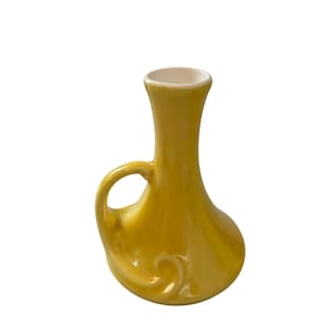 May include: A small, yellow ceramic vase with a handle and a decorative swirl design. The vase has a narrow neck and a wider base. The interior of the vase is white.