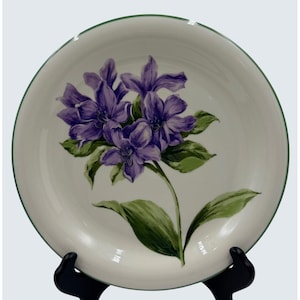 May include: A white ceramic plate with a green rim and a floral design. The design features purple flowers with green leaves and stems. The plate is displayed on a black stand.