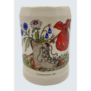 May include: Cream-colored ceramic stein with a hiking boot design. The boot holds flowers and is next to a walking stick with a red sack. The stein has the text "TuS Massenheim 1983" at the base.