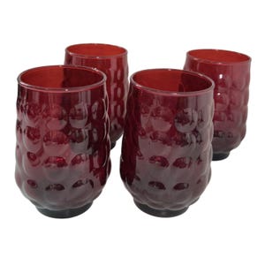 May include: A set of four deep red glass tumblers, each featuring a distinct raised circular bubble pattern across the exterior surface. These drinking glasses have a rounded, cylindrical shape with a smooth rim and a solid, weighted base.