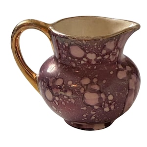 Könnte beinhalten: Ein kleines Milchkännchen aus Keramik mit einer glänzenden, irisierenden violetten Glasur und einem gesprenkelten, blasenartigen Muster. Das Kännchen hat einen bauchigen Körper, einen geschwungenen Ausguss und einen goldfarbenen Henkel.