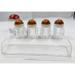 Könnte beinhalten: Ein Gewürzregal-Set aus klarem Glas mit vier kleinen, klaren, gerippten Gewürzgläsern mit weißen Deckeln und Obstmotiven. Ein größeres Glas mit Obstmotiv ist ebenfalls vorhanden. Das Set steht auf einem transparenten Kunststoffregal.