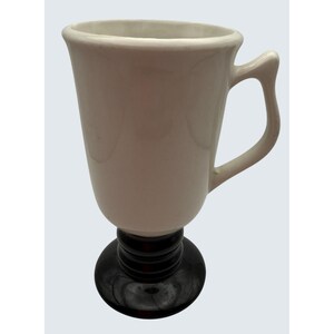 Puede incluir: Una taza de cerámica blanca con un diseño único, con un tallo y una base negra. La taza tiene un asa curva y una abertura ancha. La taza está diseñada para bebidas.