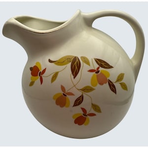 May include: Cream-colored ceramic pitcher with a rounded body and a curved handle. The pitcher is decorated with a floral pattern in shades of yellow, orange, and brown. The pitcher has a spout for easy pouring and is suitable for serving beverages.