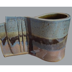 May include: A handmade ceramic mug with a unique, flowing design. The mug features a blend of colors, including blue, brown, and tan, with a textured surface. The handle is integrated into the body, creating a seamless look. The mug is ideal for coffee or tea.