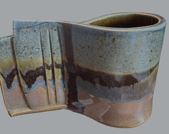 May include: A handmade ceramic mug with a unique, flowing design. The mug features a blend of colors, including blue, brown, and tan, with a textured surface. The handle is integrated into the body, creating a seamless look. The mug is ideal for coffee or tea.