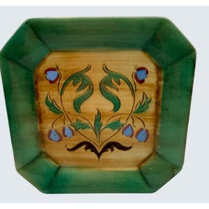 May include: A square, green-edged decorative plate with a floral design. The center of the plate is tan with a symmetrical pattern of green leaves, blue flowers, and red accents. The plate has an octagonal shape.
