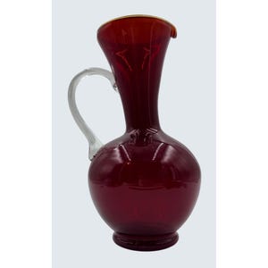 May include: A ruby red glass pitcher with a clear glass handle. The pitcher has a bulbous base and a slender neck with a gold rim. The handle is curved and attached to the side of the pitcher. A decorative piece for serving beverages.