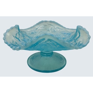 May include: A light blue, vintage glass centerpiece bowl. The bowl has a ruffled edge and is supported by a round base. The glass is decorated with a leaf pattern. The bowl is likely used for serving fruit or displaying decorative items.