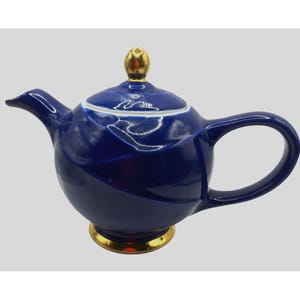 May include: A navy blue ceramic teapot with a rounded body, a curved spout, and a large handle. The lid is topped with a gold-coloured knob. The base and the rim of the teapot are also gold-coloured. The teapot has a white and blue swirl design.